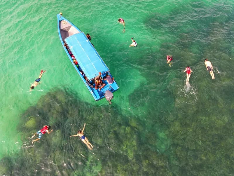 Aerial view of people standing and swimming in waist-deep clear water around an anchored boat
